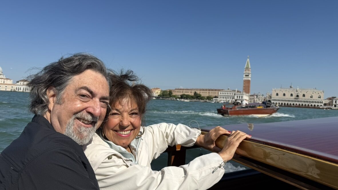 Two friends smiling in the back of a water taxi passing the San Marco waterfront with the bell tower behind them