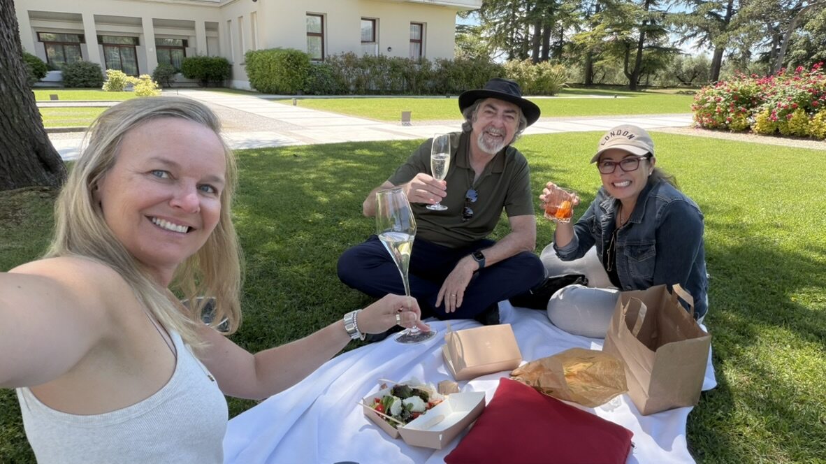 She-Stache grinning in a selfie with friends sitting behind her on a picnic blanket