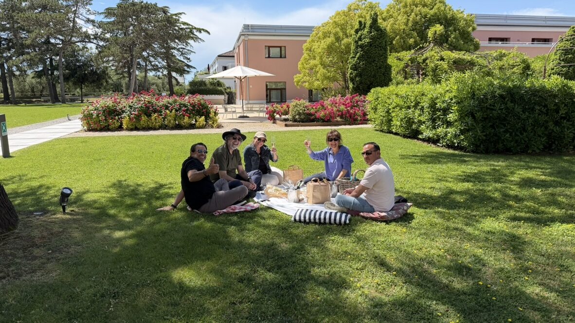 The group of friends having a picnic on blankets on a wide green lawn at the JW Marriott Venice, hotel buildings and trees behind