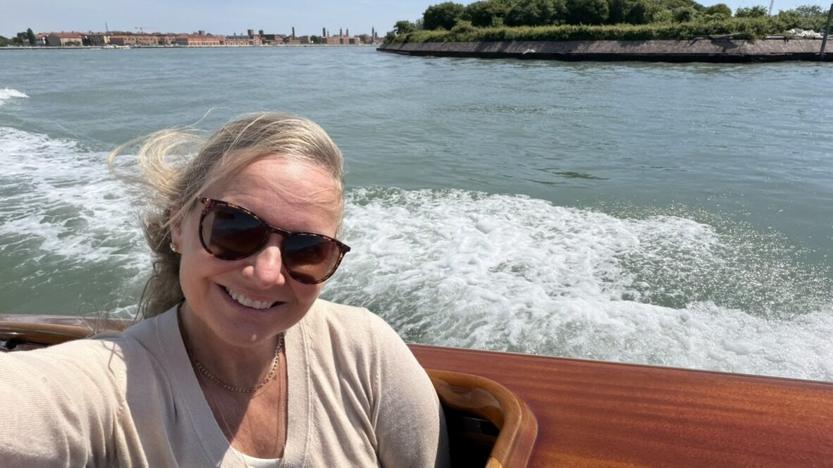 She-Stache smiling in sunglasses at the back of a speeding water taxi, wake spraying white across the lagoon